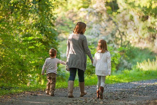 Eine Frau geht auf einem Waldweg mit ihrer ungefähr achtjährigen Tochter und dem vierjährigen Sohn. In einer entspannten, aber stillen Atmosphäre. Die drei Figuren sind von hinten fotografiert.